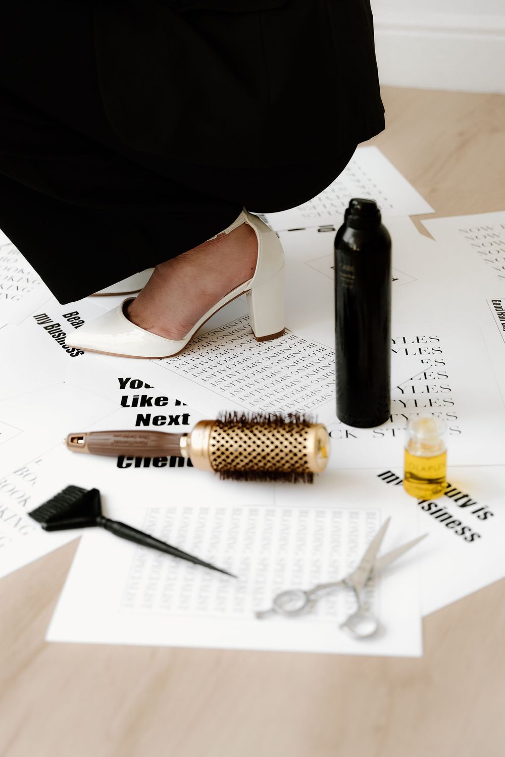 Hairdressing tools including scissors, round brush and hairspray next to a white high heel shoe on white paper.