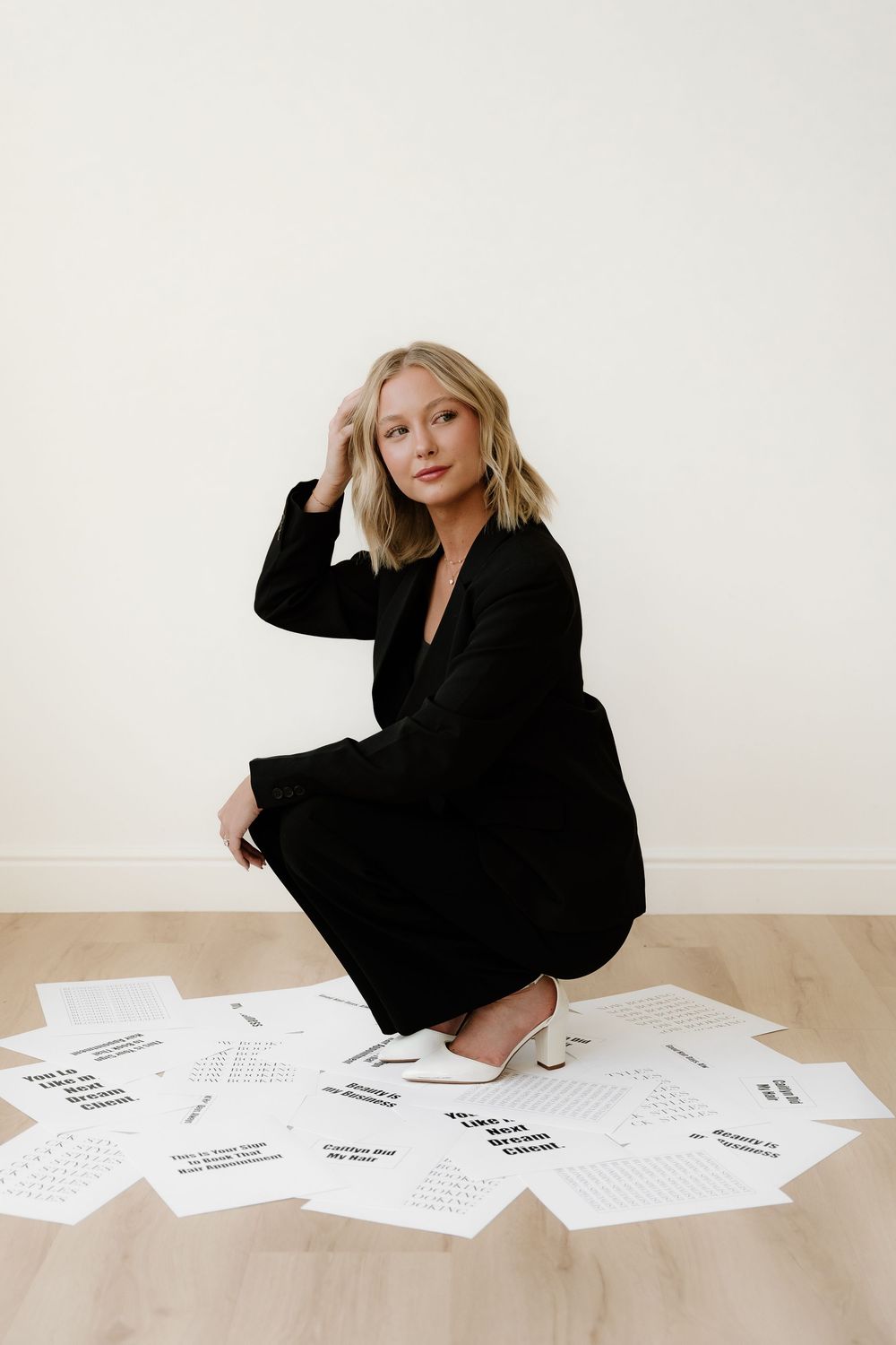 Stylish figure in black outfit sitting among scattered papers on hardwood floor.