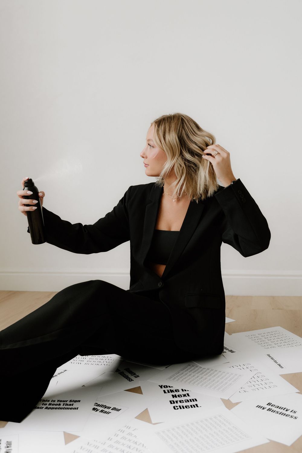 Person in black blazer styling hair while sitting among scattered papers on floor.