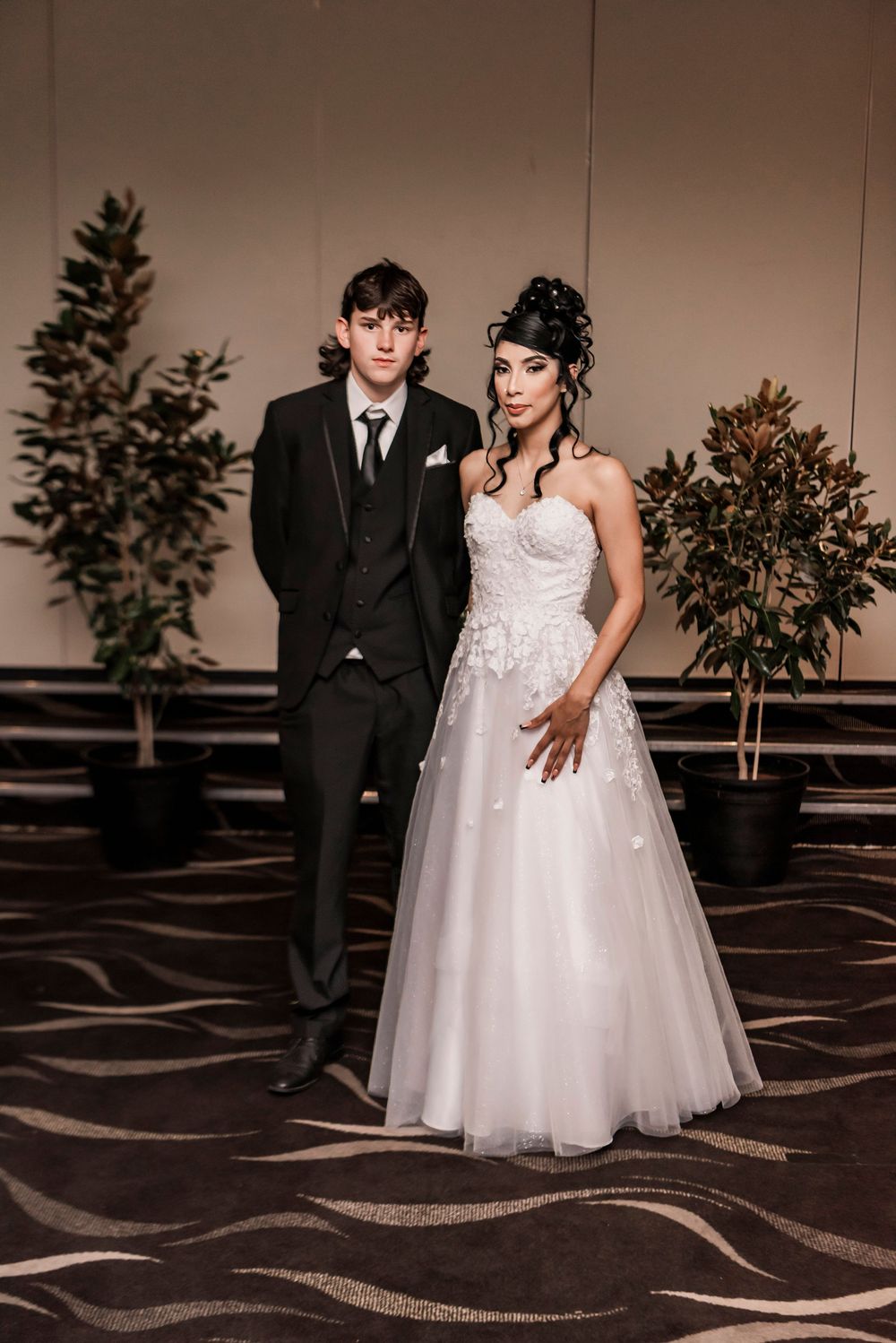 A couple stands elegantly in a ballroom with a dark carpet and potted plants, dressed in formal wedding attire.