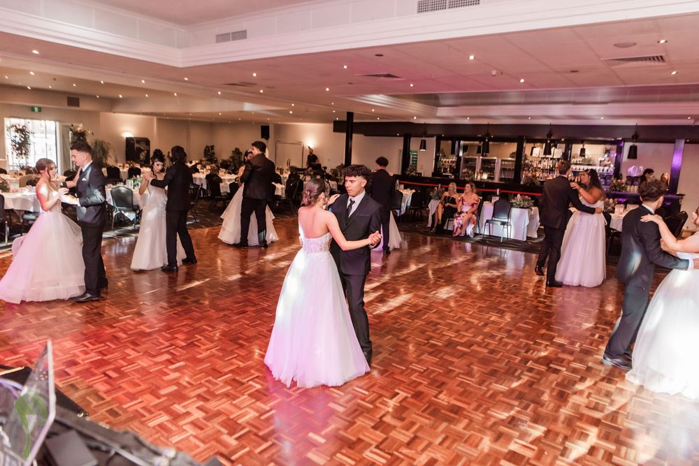 Wedding guests dance and celebrate on a large ballroom floor with pink lighting and white decorations.