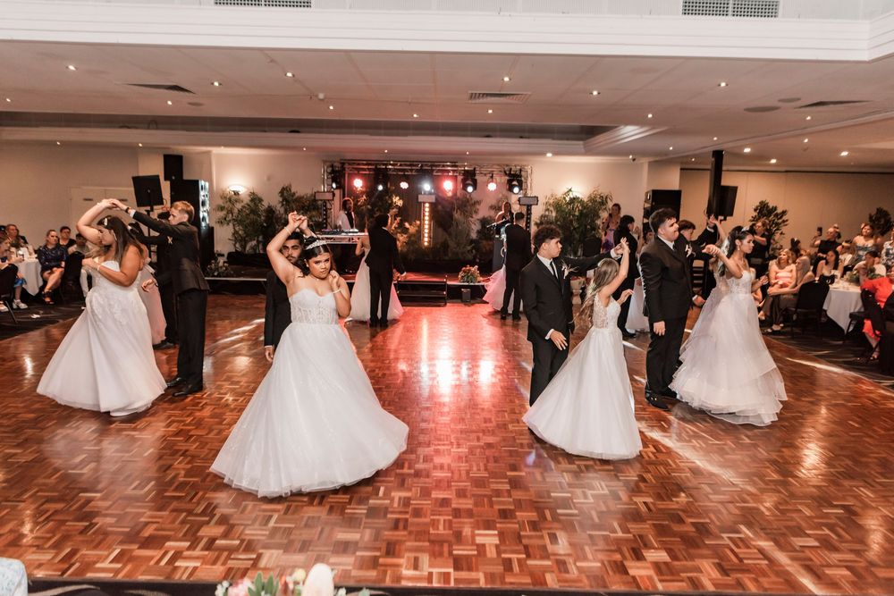 Several couples dance together in elegant evening attire on a parquet dance floor under warm lighting at a formal event.