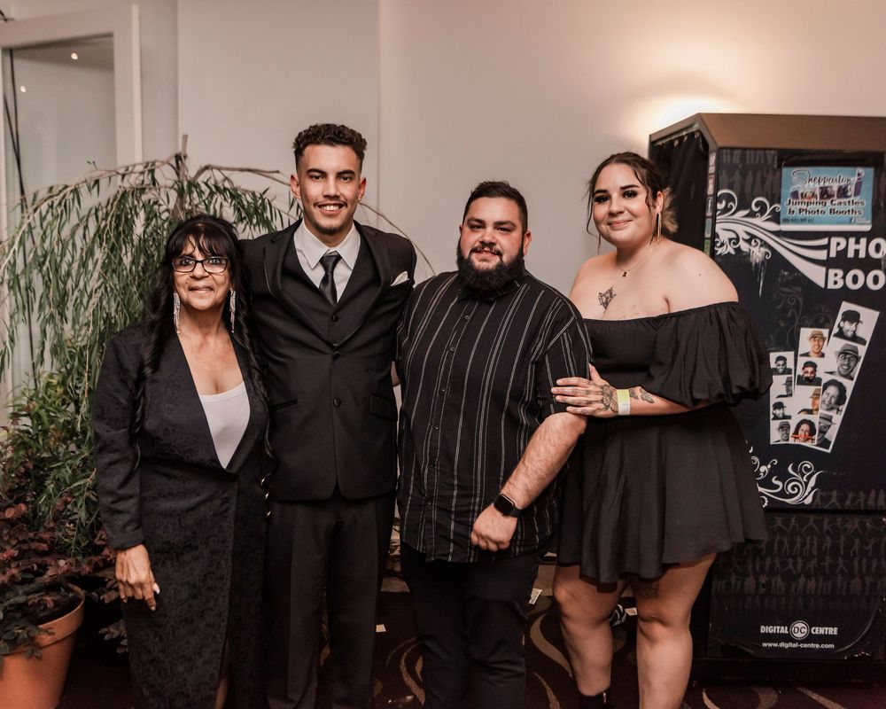 A group of four people in formal black attire pose together at an event with a decorative plant in the background.
