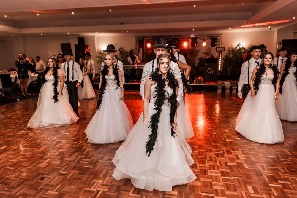 Young debutantes in white ball gowns dance elegantly across a shining parquet floor during a formal gathering.