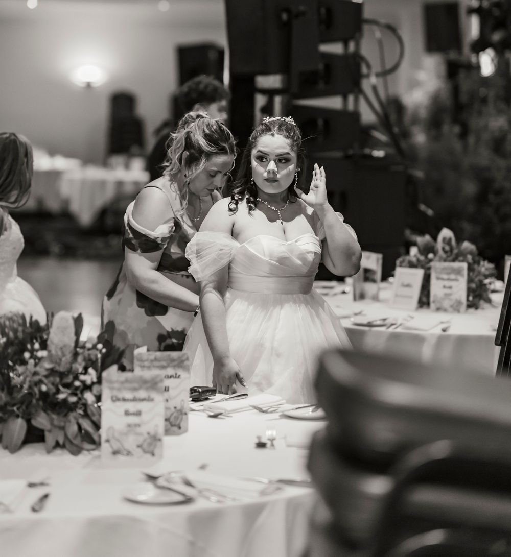 A bride in a white dress gestures while seated at an elegant reception table with floral centerpieces.