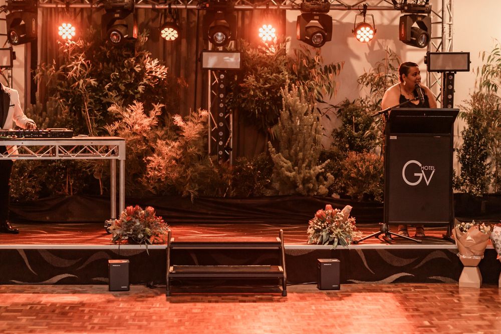 DJ booth setup at an event with warm lighting, wooden backdrop, floral arrangements and stage lighting.