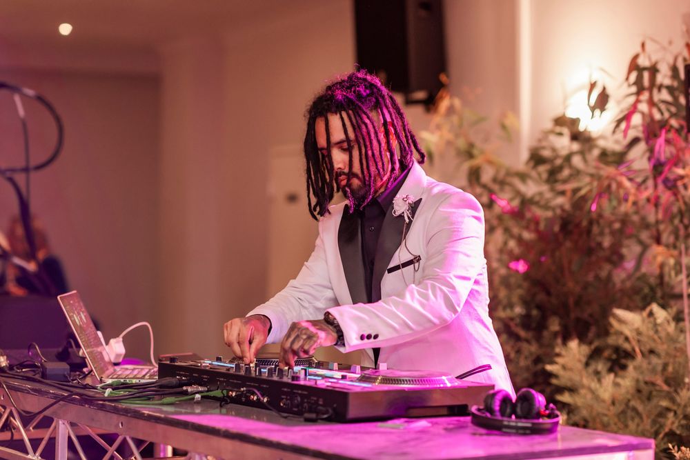 DJ performs with equipment under pink lighting at an event with floral decorations in the background.