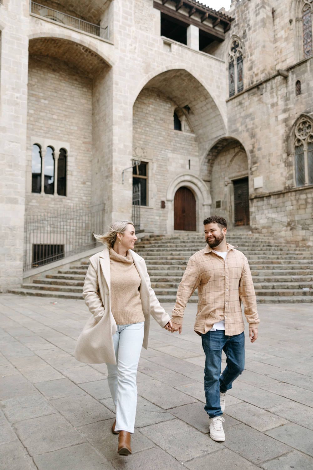 Couple walking hand in hand through a historic stone courtyard with grand staircase.