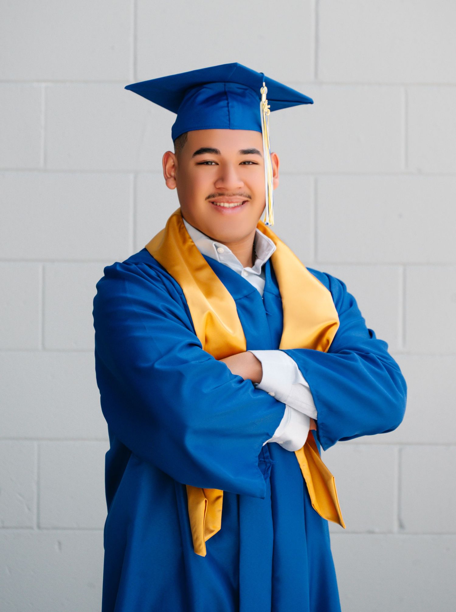 A graduate wearing traditional blue cap and gown with gold stole poses against a white brick wall in three poses.