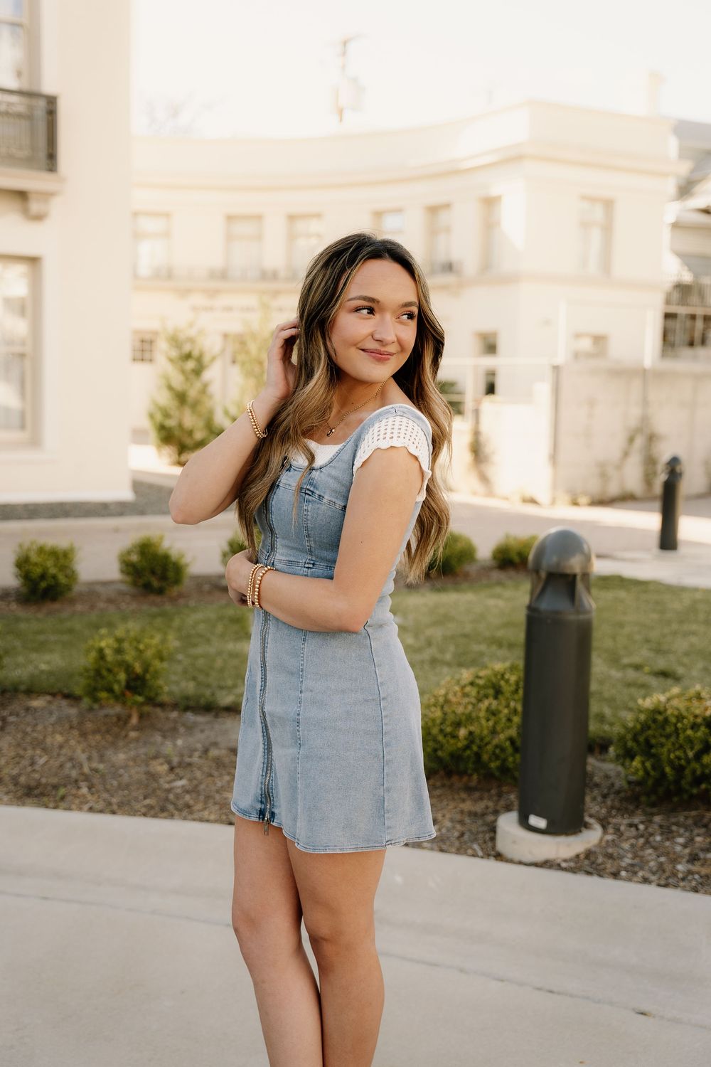 Light denim overall dress styled with white top in elegant outdoor setting.