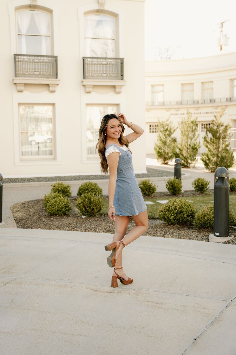 A stylish summer outfit with denim dress and brown sandals photographed at sunset by historic building.