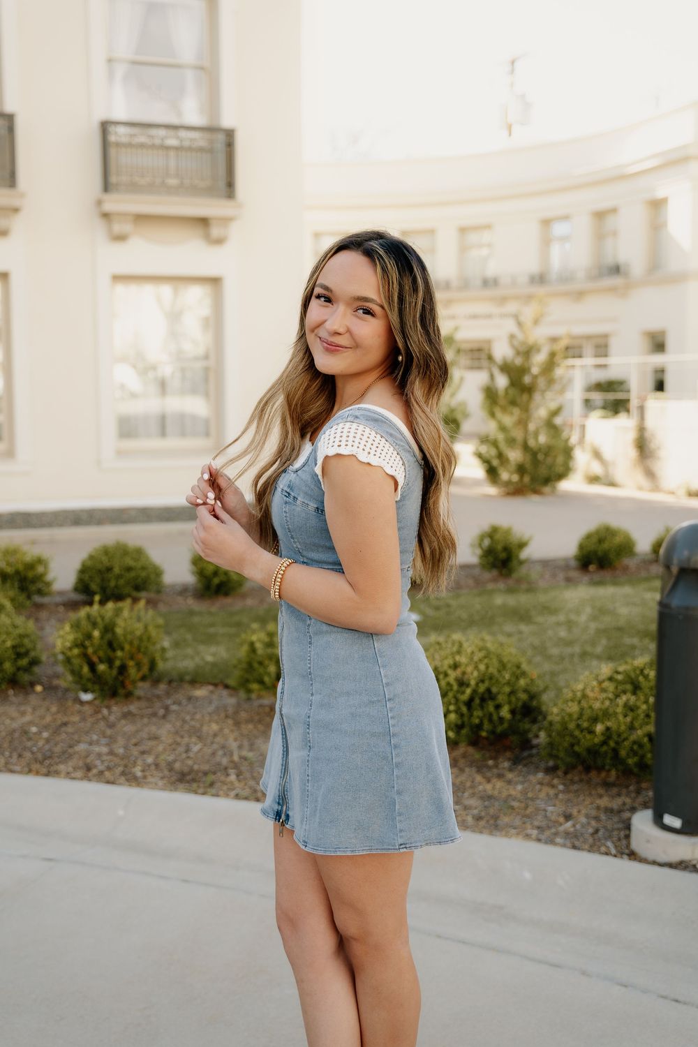 Woman in denim dress and white top poses outdoors near cream colored building with landscaping.