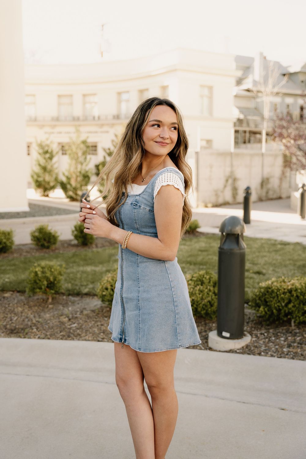 A casual spring fashion look with denim overall dress against landscaped building grounds.