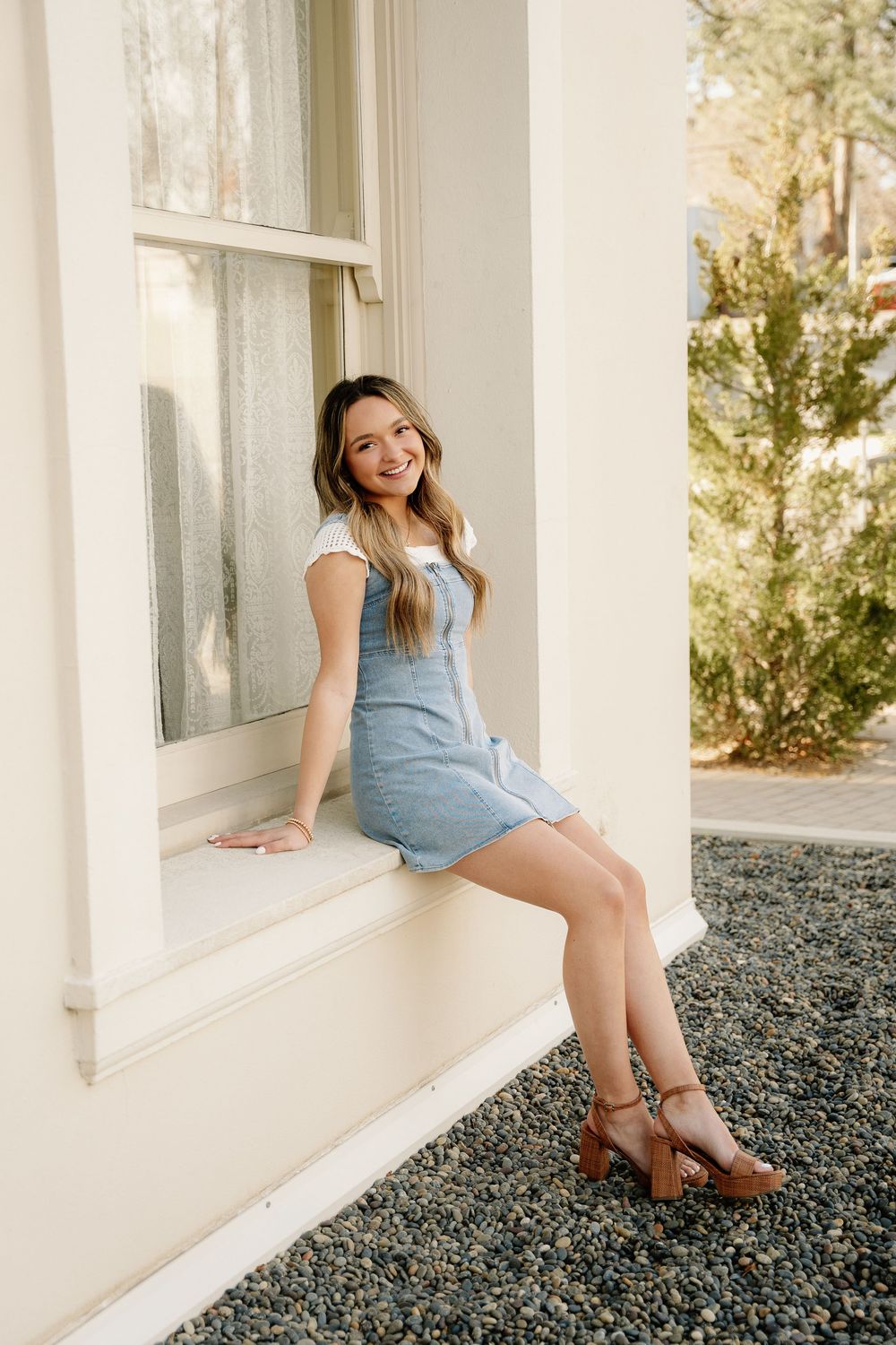 Casual portrait on window ledge wearing light denim dress and brown platform sandals.
