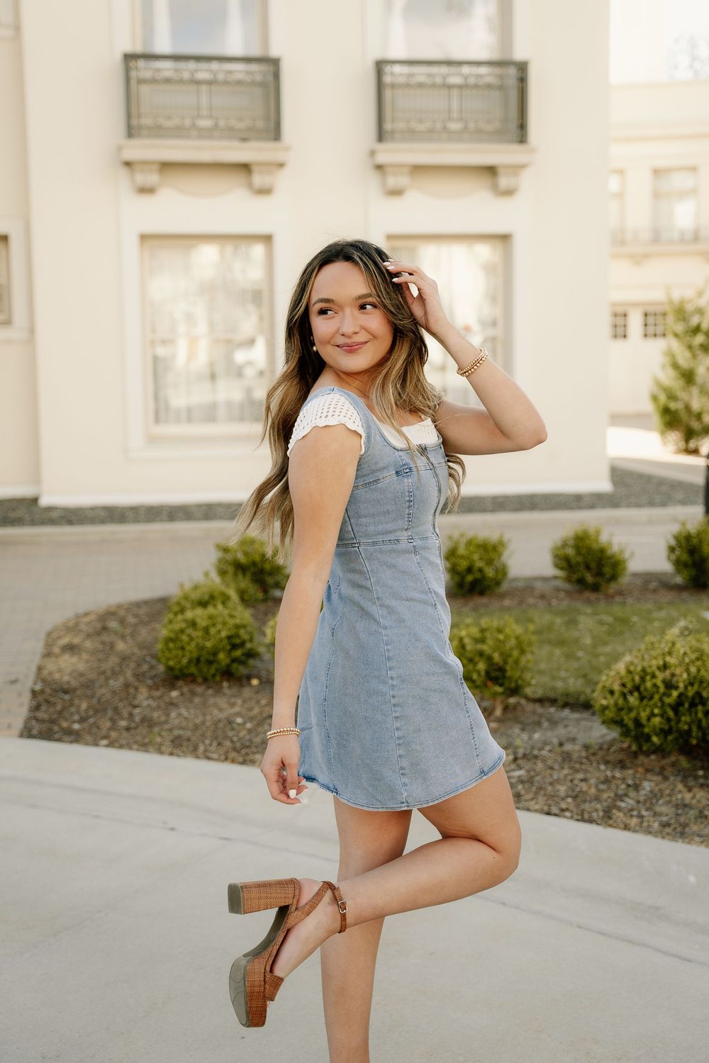 Stylish denim overall dress with brown heels shown in playful outdoor fashion pose against building exterior.