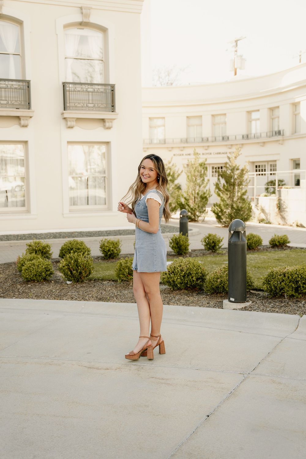 Casual denim dress outfit photographed against curved architectural backdrop with manicured garden.