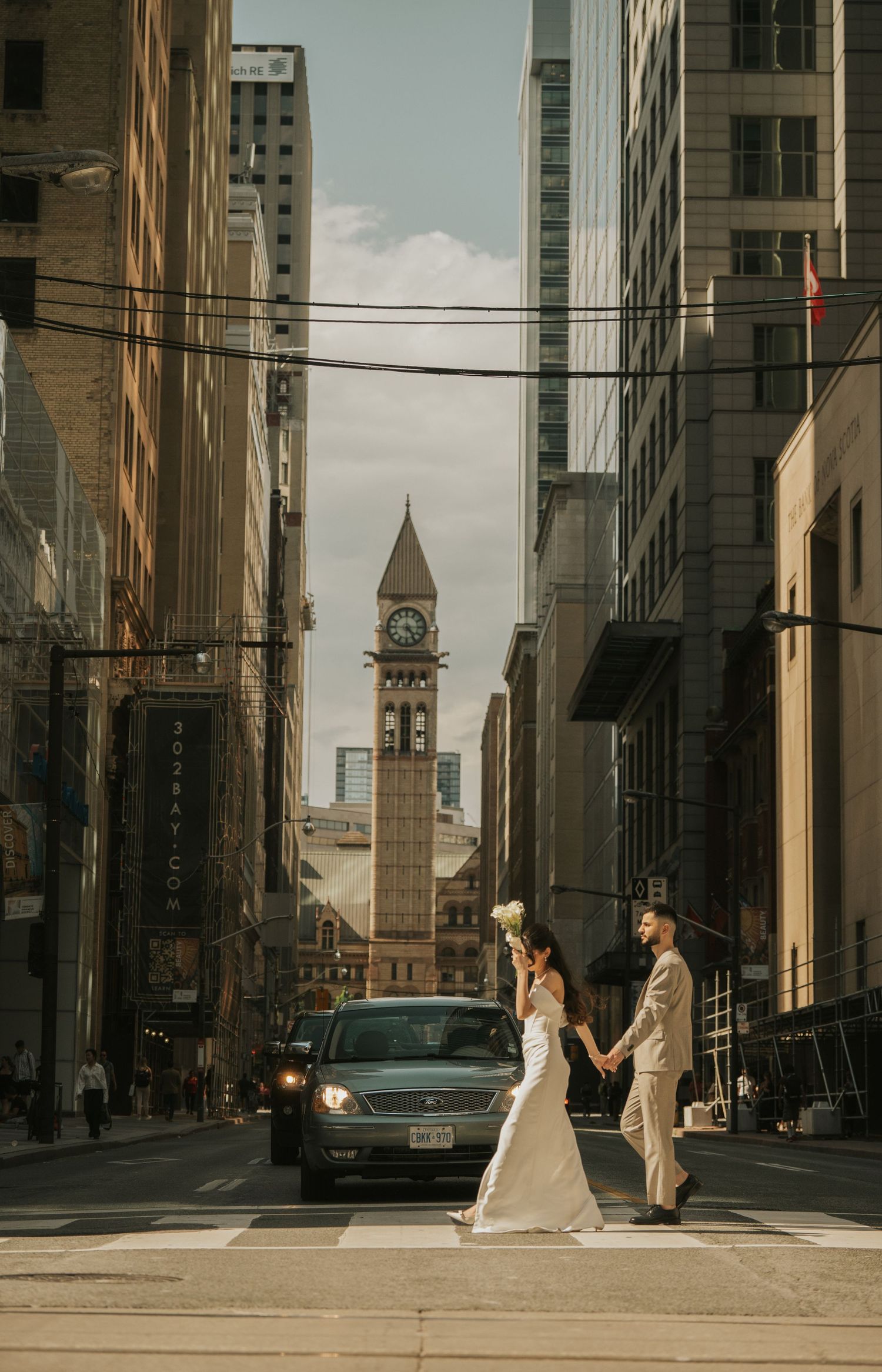 Newlywed couple crossing urban street with historic clocktower and tall buildings in downtown cityscape background.