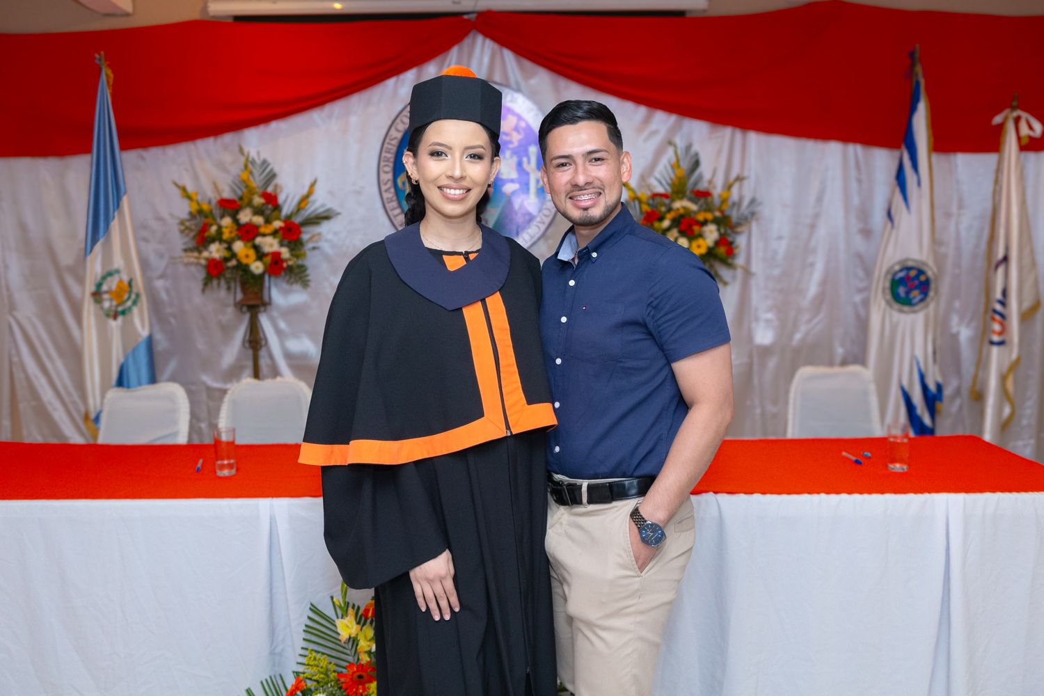 Two people pose together at a formal event with red and white decorative elements in the background.