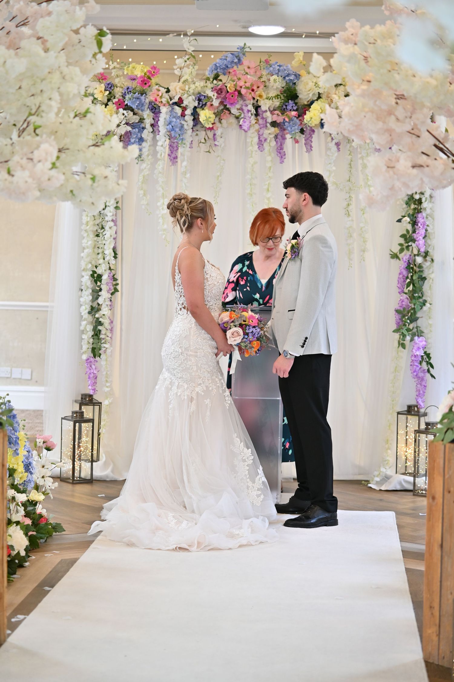 A wedding ceremony takes place under a floral arch with delicate hanging decorations and soft lighting.