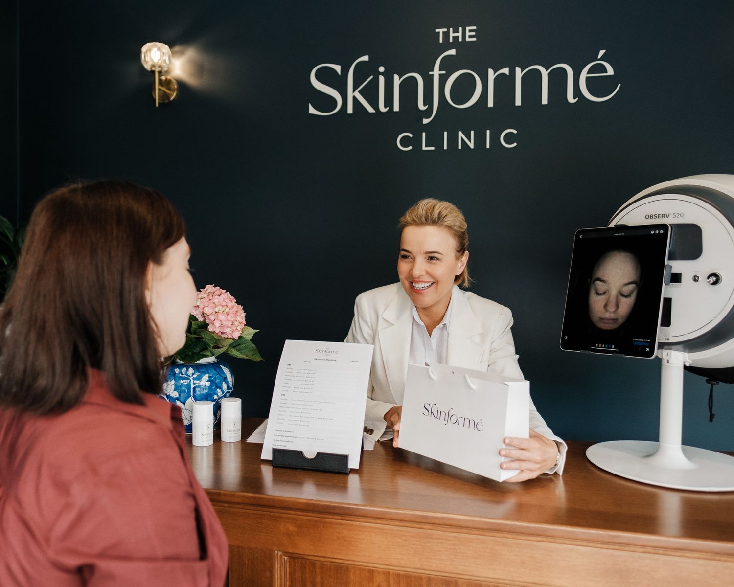 A medical professional in a white coat greets a client at the reception desk of The Skinforme Clinic.