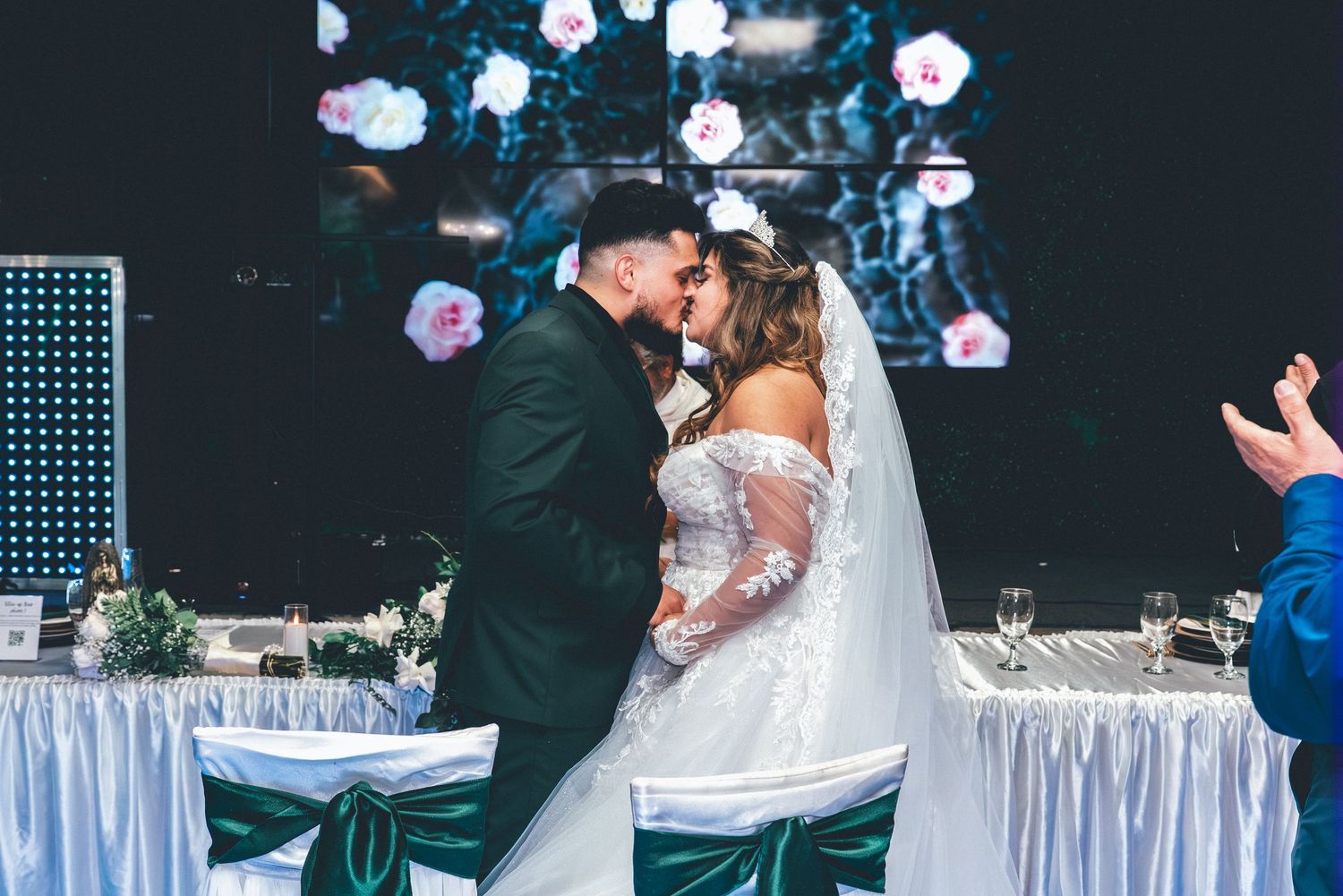 A romantic wedding moment captured at a reception with white draped tables and sparkling lights in the background.