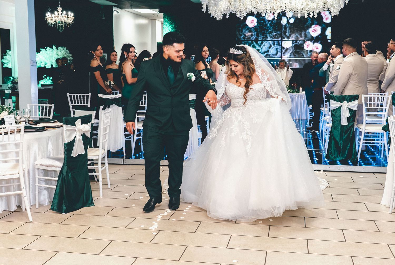 Wedding celebration hall decorated with white linens and chandeliers showing guests mingling at tables.