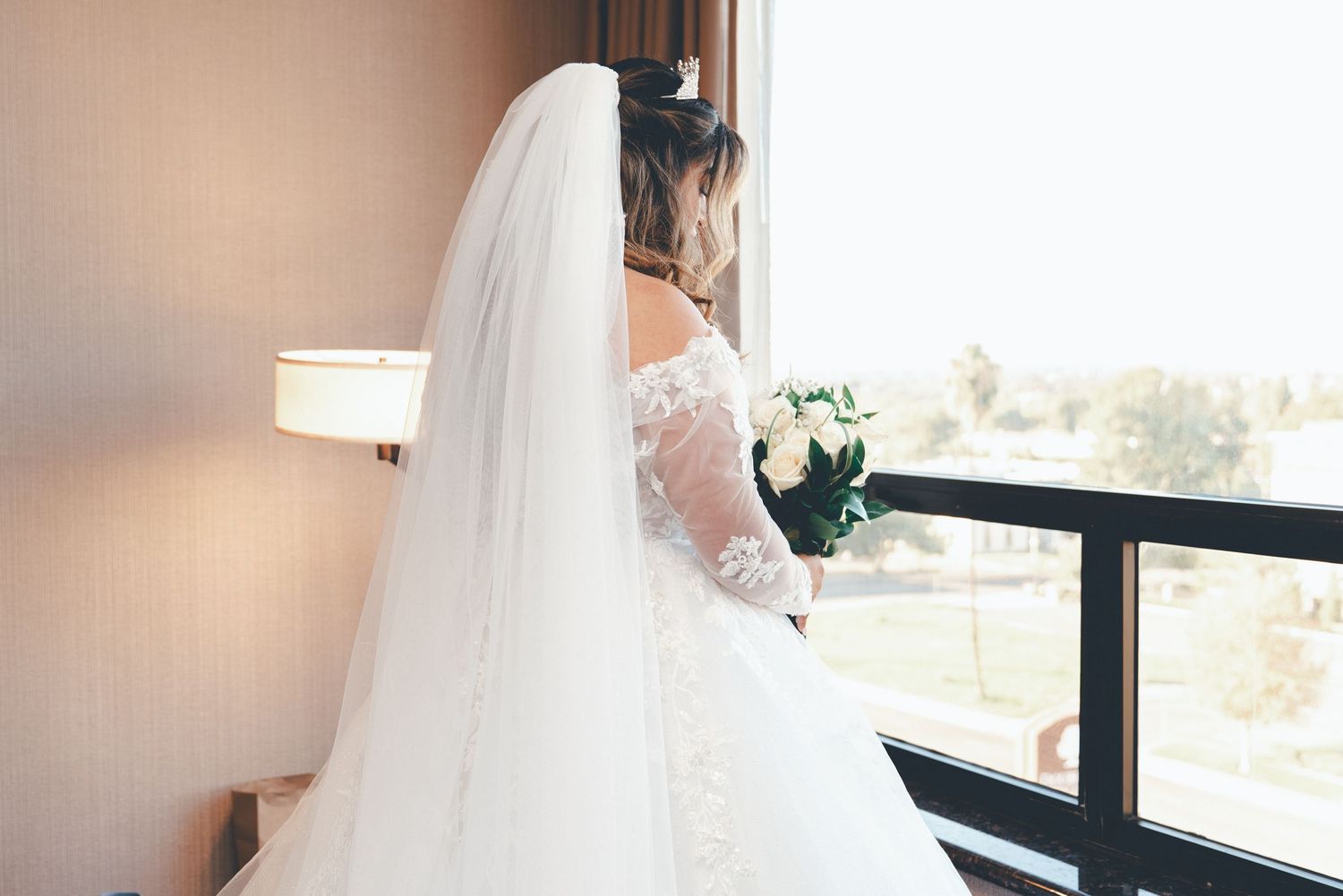 A bride in a white wedding dress adjusts her veil while getting ready in a sunlit room.
