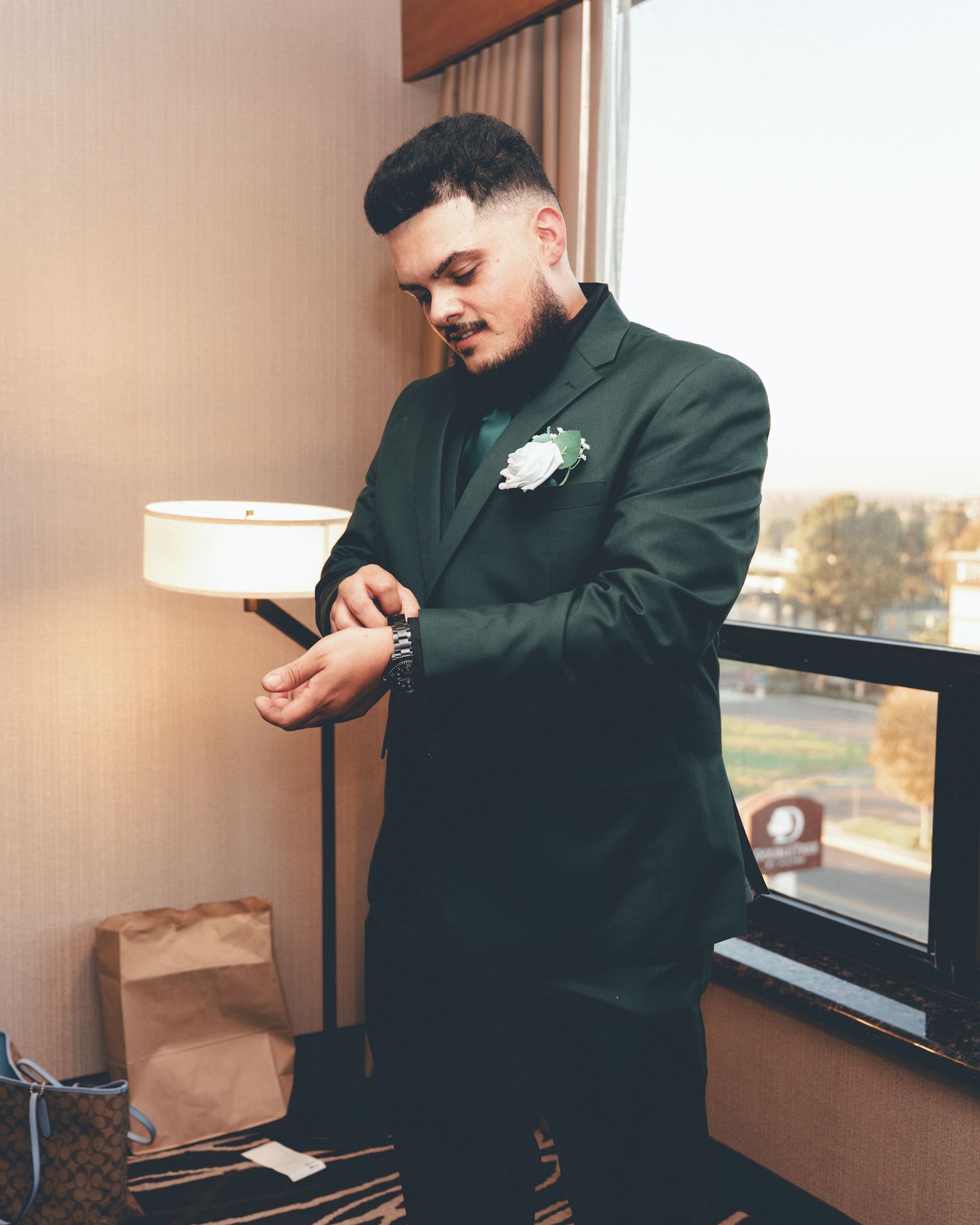 A person in a dark suit and boutonniere checks their watch while standing in a room with paper bags and a window.