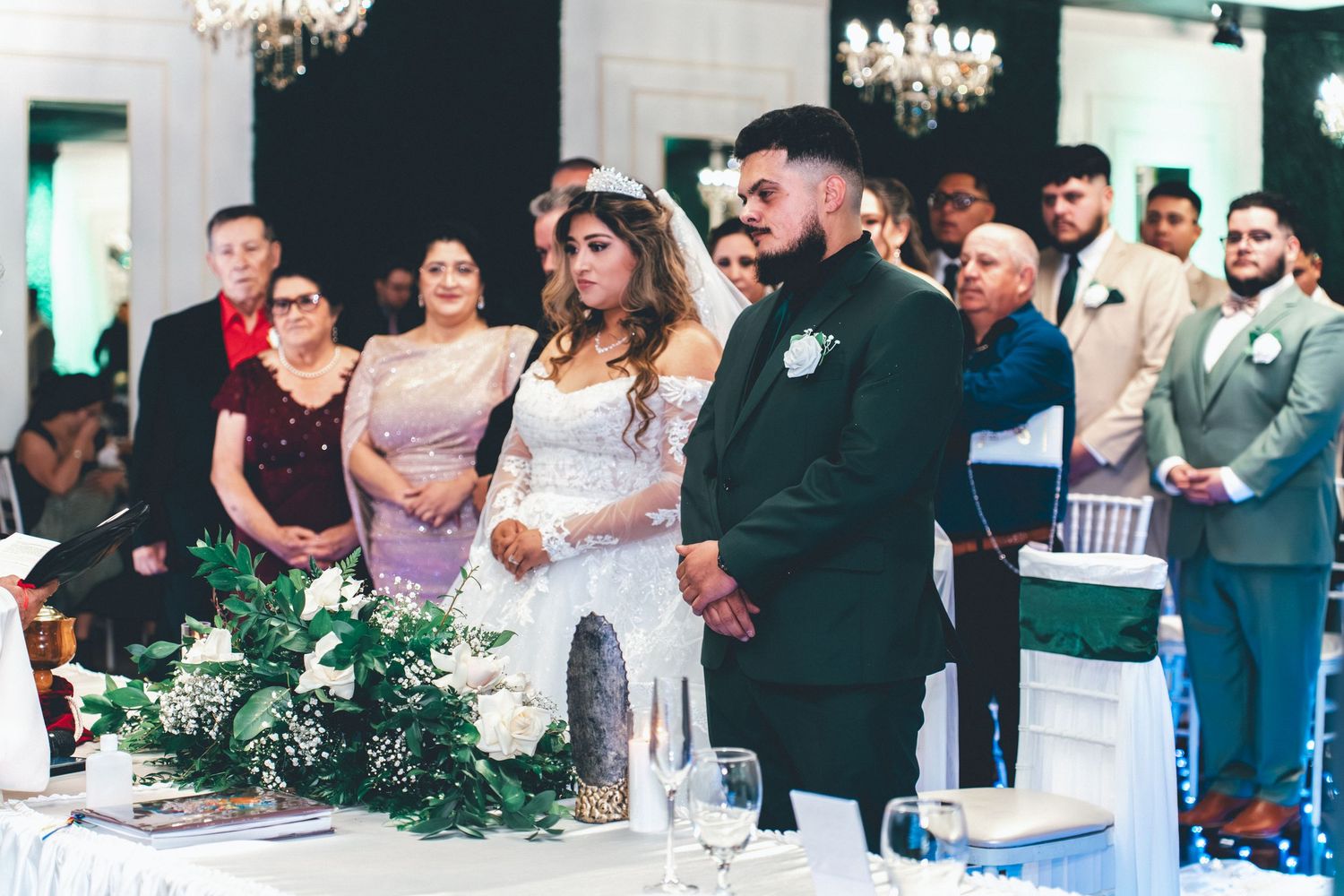 Wedding guests gather around tables at an elegant reception with chandeliers and white decor.