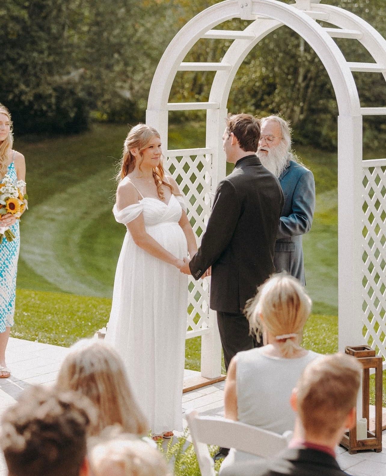 A wedding ceremony takes place under a white lattice archway in an outdoor garden setting.