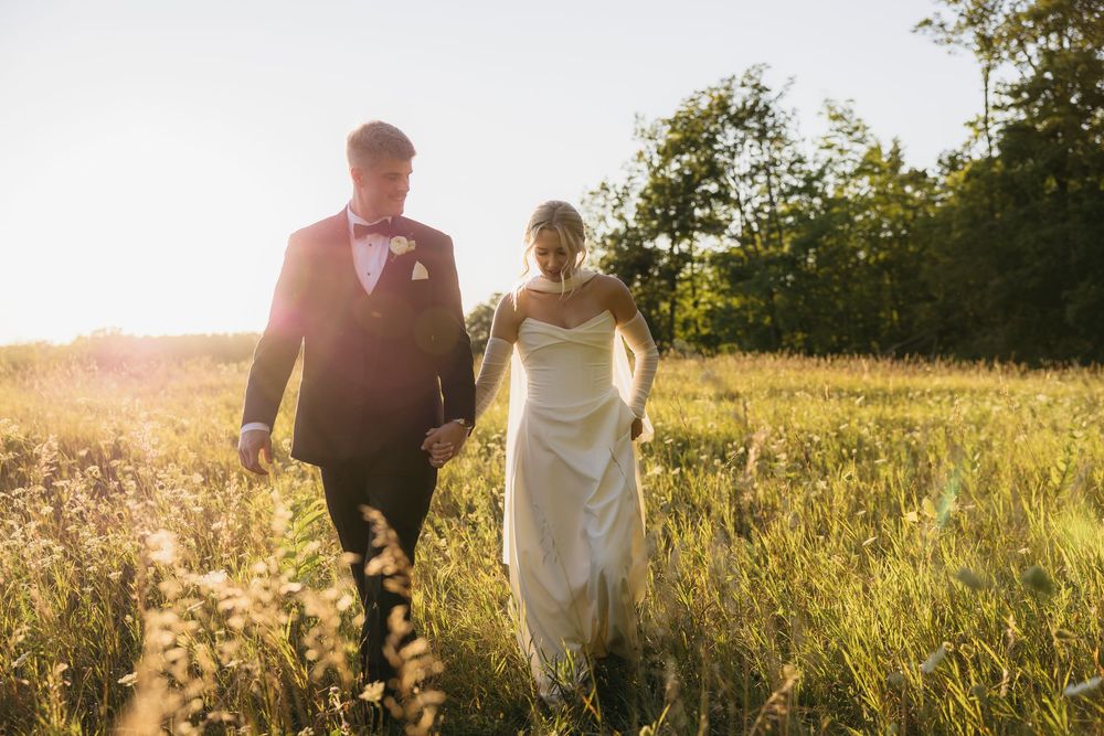 Newlyweds walking hand in hand through a sun-drenched field at sunset wearing formal wedding attire.
