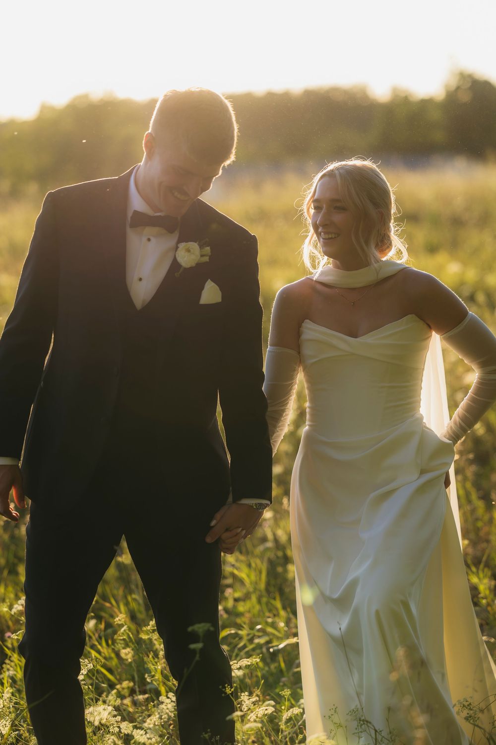 A couple in wedding attire stands together in a golden sunlit field during sunset.