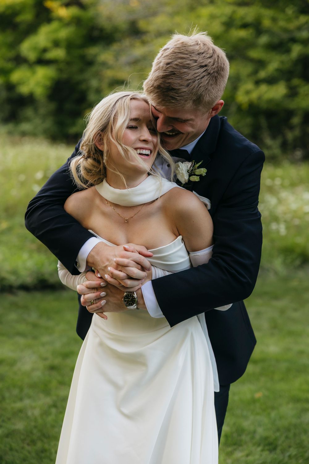 A couple in wedding attire embrace and laugh joyfully together in a lush green outdoor garden setting.