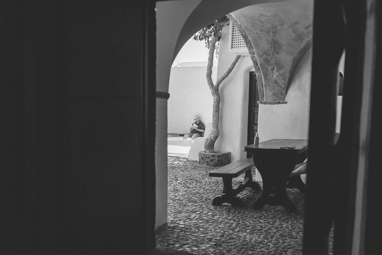 Black and white photograph showing through an arched doorway with a man sitting in a courtyard n the distance by a pool.