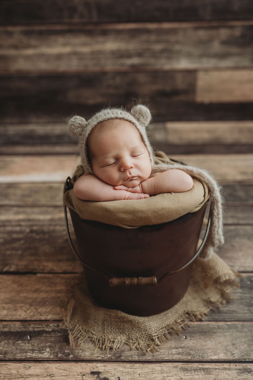 Sleeping newborn wearing bear ears bonnet nestled in vintage wooden bucket on rustic floor.