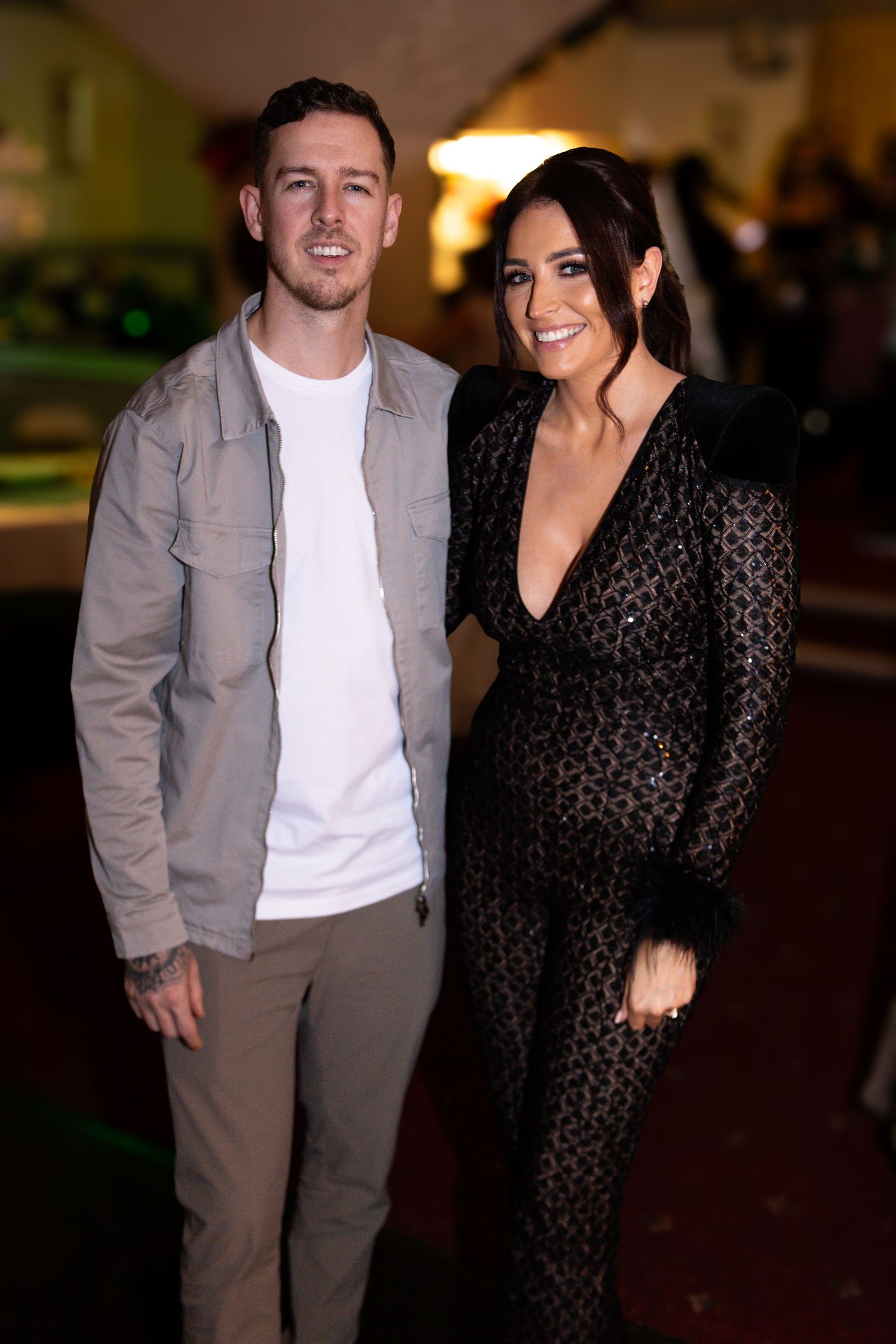 A couple poses together at an event, with one wearing a casual jacket and white shirt and the other in a black patterned jumpsuit.