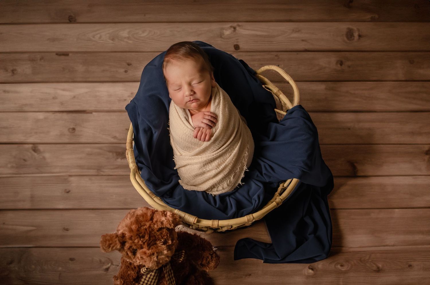 Newborn portrait photography of a baby boy wrapped in neutral fabric and nestled in a basket on a wooden floor.