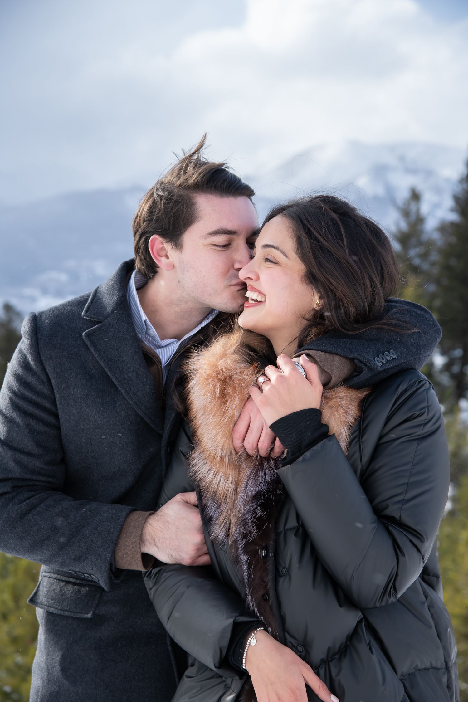 A couple in winter coats shares a romantic moment outdoors with snow-capped mountains in the background for Breckenridge engagement photos.