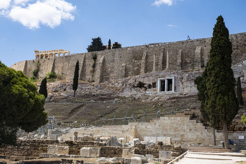 Ancient stone walls and cypress trees line the archaeological ruins of a historic Mediterranean site in panoramic view.