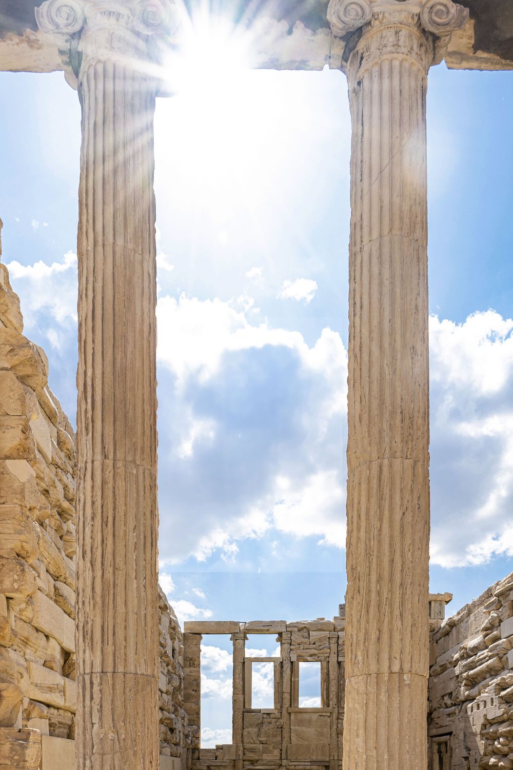 Ancient Greek temple columns rise against a bright blue sky with sunlight streaming through the Corinthian capitals.