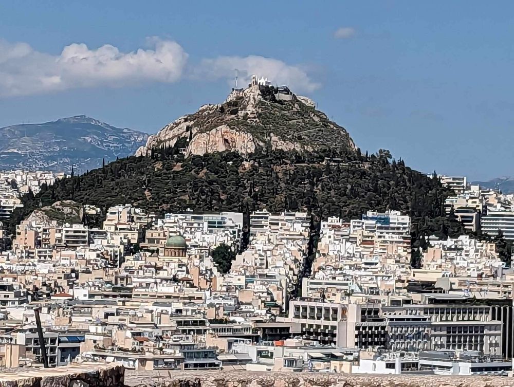 Panoramic cityscape view of Athens with densely packed white buildings climbing up hillsides under blue skies.