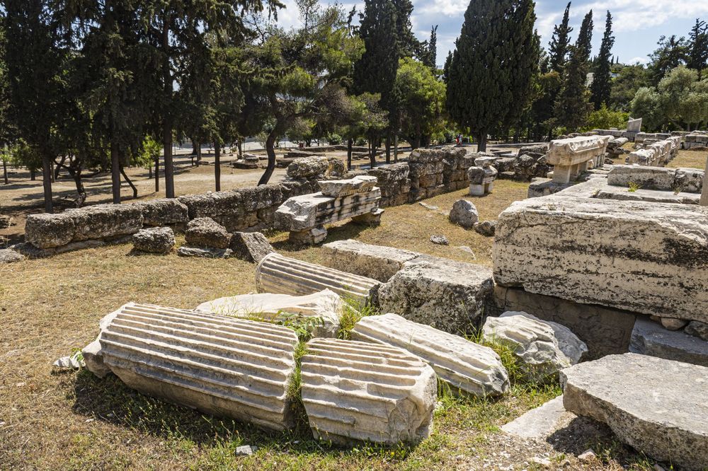 Archaeological ruins with fallen stone columns and blocks scattered among cypress trees in an ancient Greek site.