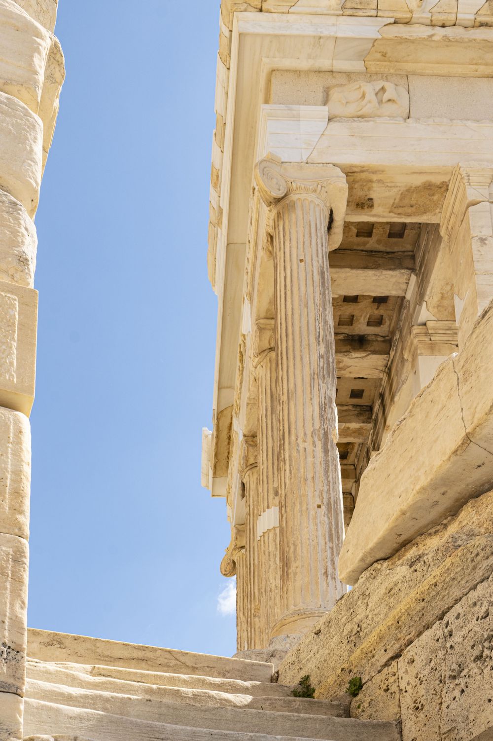 Ancient Greek temple columns and architectural details of the Acropolis against a bright blue sky in Athens, Greece.