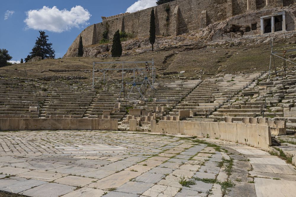 Ancient stone amphitheater ruins with rows of weathered seating and architectural details against a blue cloudy sky.