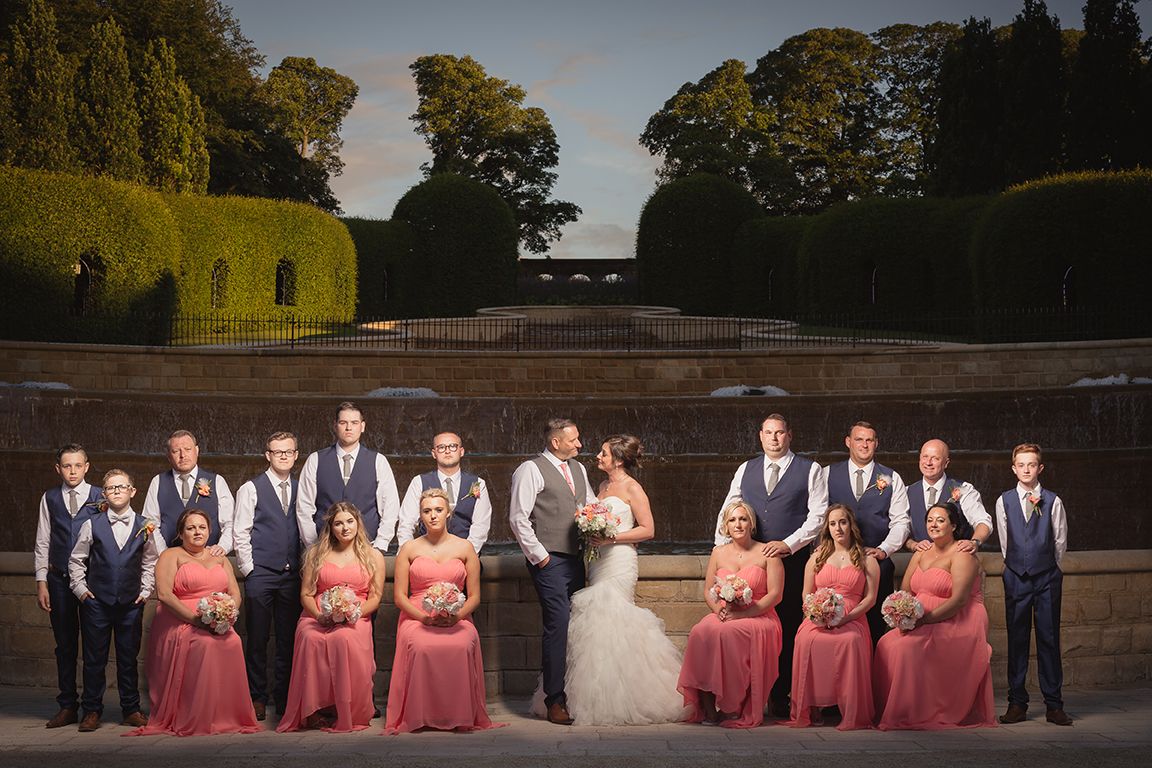 Wedding party poses formally in a garden with bridesmaids in coral dresses and groomsmen in gray vests at sunset.