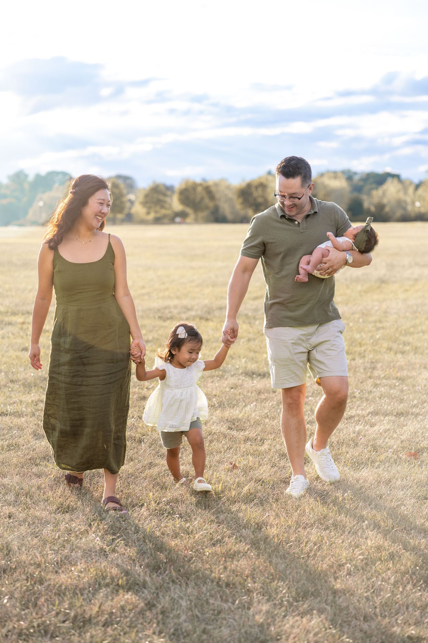 Family walking together holding hands in a golden field during sunset with tall grass and trees in background.