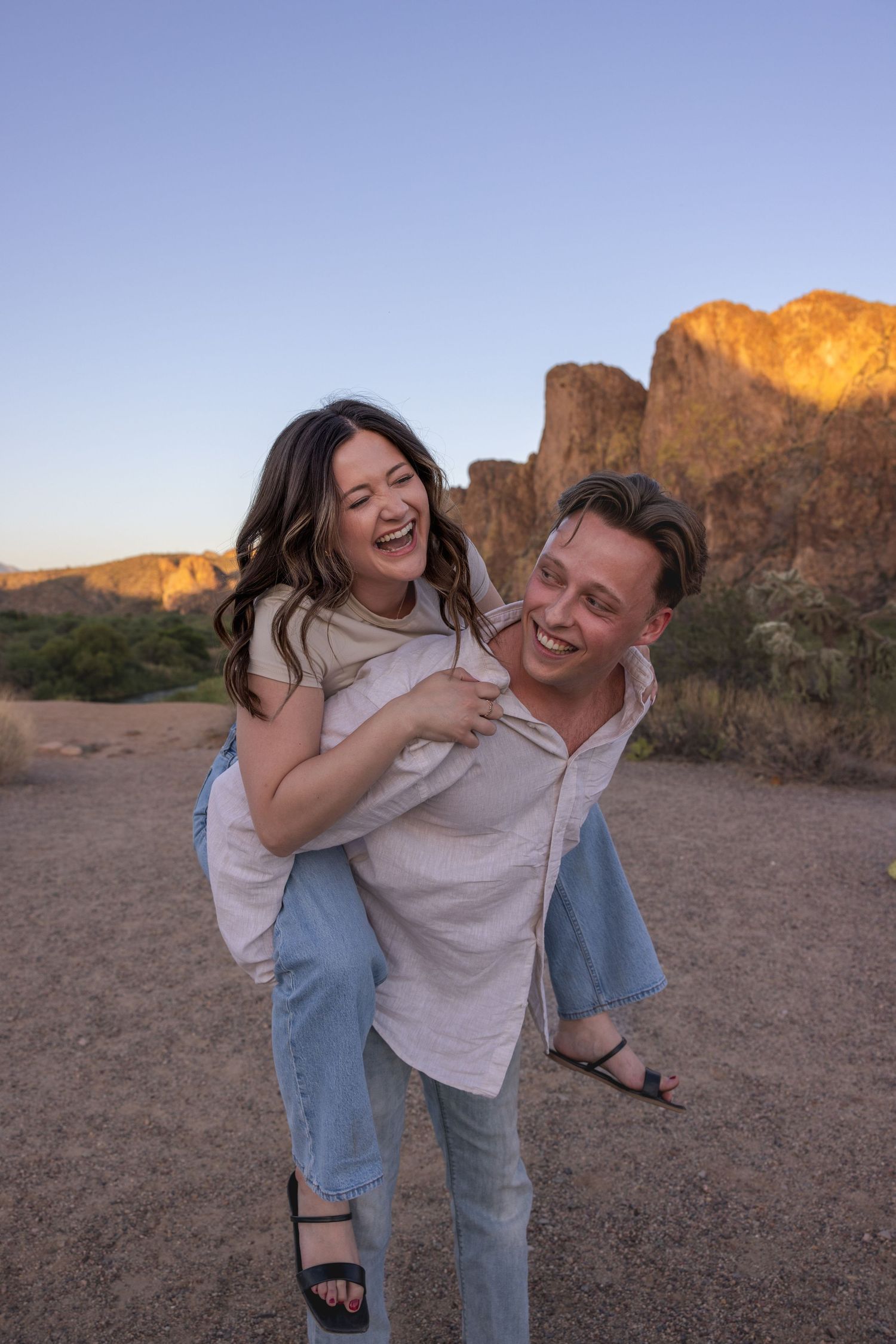 Laughing couple shares a joyful piggyback ride in the Arizona desert at sunset with glowing red rock mountains behind them.