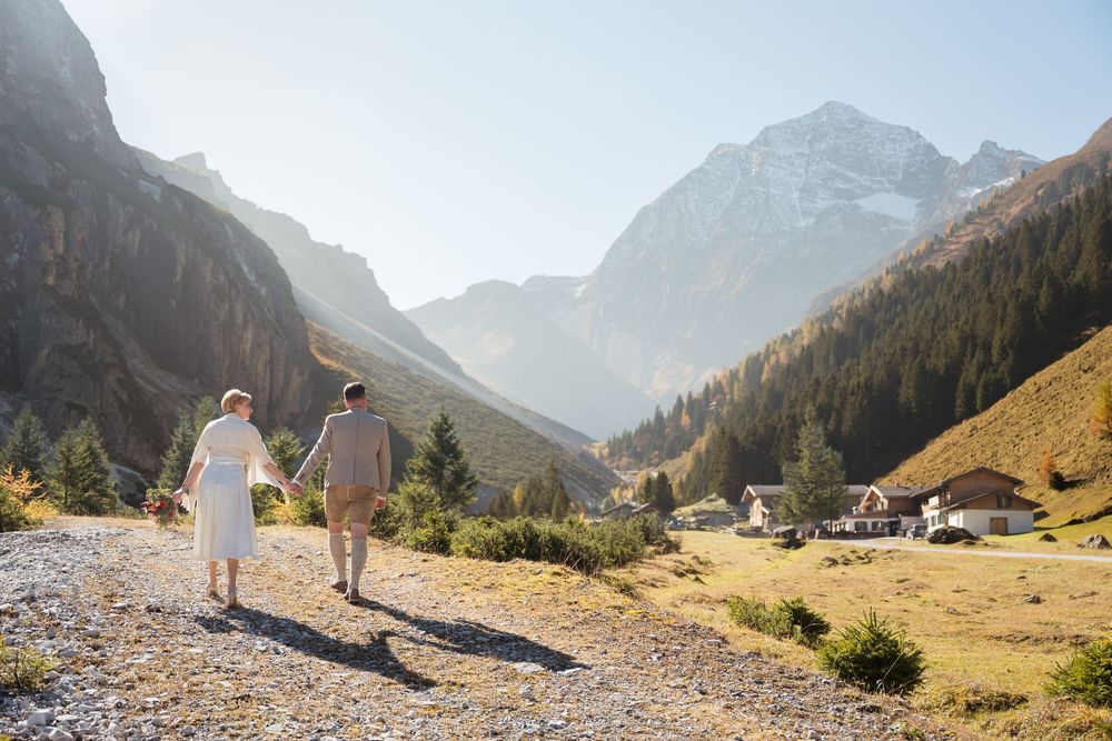 Brautpaar wandert Hand in Hand durchs Stubaital, im Hintergrund ein Bergdorf vor schneebedecktem Gipfel