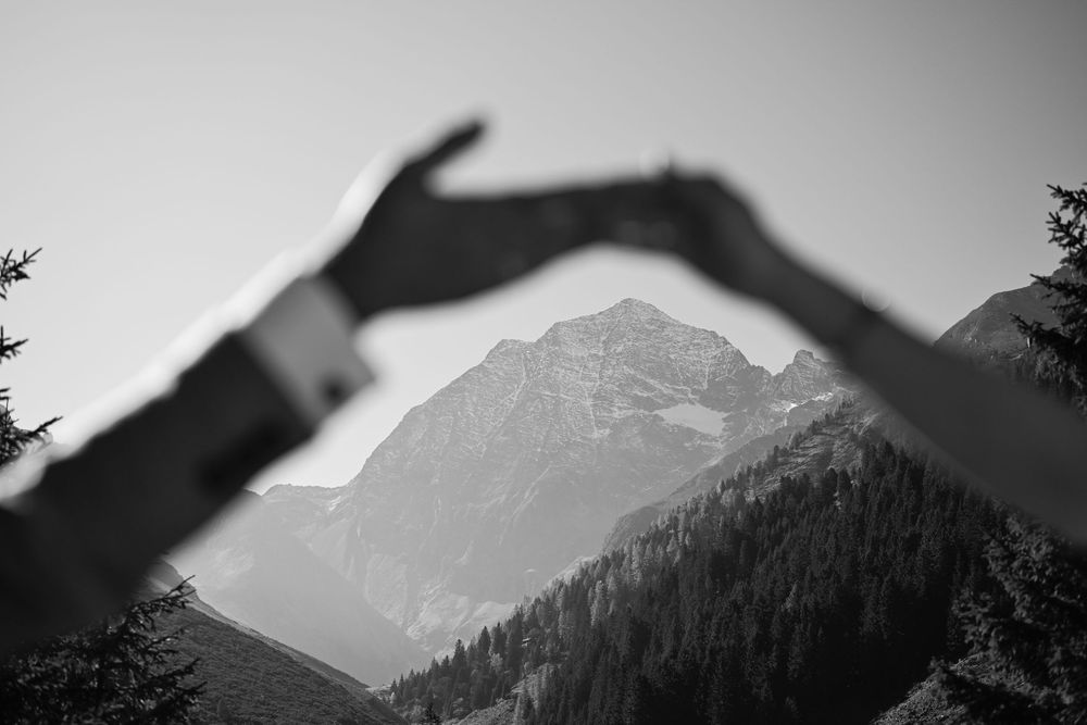 Schwarz-Weiß-Foto: Hände formen ein Herz vor der Bergkulisse Tirols