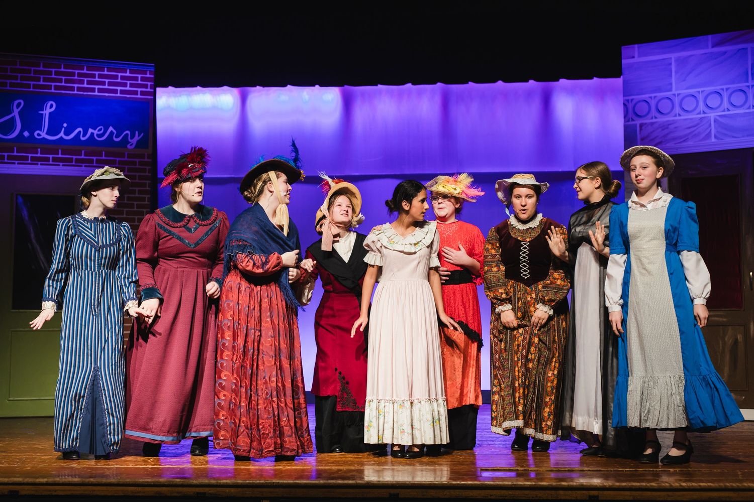 Performers in period costumes dance and interact on a colorfully lit stage during a theatrical production.